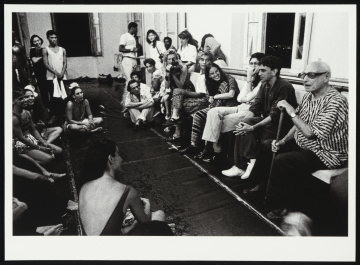 Fotografia de sala que aparenta ser destinada para ensaios com pessoas sentadas e em pé. A maioria está voltada para Pierre Verger, que está sentado segurando bengala. Caetano Veloso está ao lado de Verger.