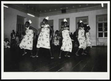 Mulheres vestidas com vestidos brancos com estampas de folhas. Elas usam colares e brincos e ornamentos brancos na cabeça, e parecem ter um tipo de pó branco nos ombros e na região da clavícula. Elas estão posicionadas em círculo, têm as bocas abertas, e estão com as mãos próximas à cintura, no que parece ser um movimento de dança. Ao fundo é possível ver a parede com janelas.