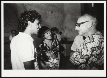 Caetano Veloso, Pierre Verger e mulher não identificada parados em pé olhando uns para os outros, enquanto sorriem, aparentemente conversando.