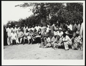 Fileira de pessoas sentadas (Gilberto Gil está entre elas), algumas delas usam óculos escuros. Atrás delas, há multidão de pessoas em pé. Na parte superior da fotografia, é possível ver copas de árvores.