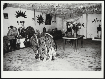 Fotografia de sala com ornamentos e ferramentas de orixás pendurados nas paredes. Há um vaso com flores em cima de uma mesa. No centro, há tecidos sobrepostos em estrutura. Do lado esquerdo da fotografia, é possível ver Pierre Verger curvado olhando para baixo.